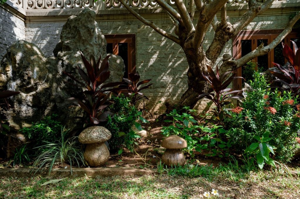 Two mushroom-shaped garden ornaments sit amongst lush greenery and rocks in front of a stone building with wooden windows.
