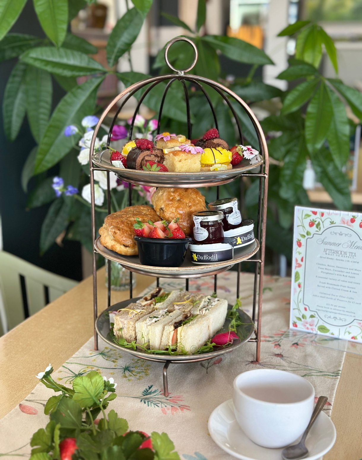 A three-tiered stand holds an assortment of afternoon tea items, including sandwiches, scones with jam and cream, and small cakes, with a cup of tea and a menu nearby.